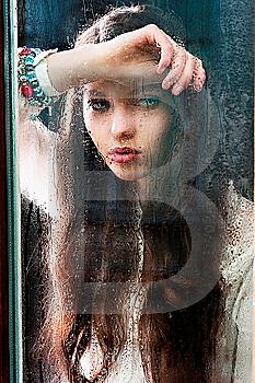 Portrait of a lovely young lady looking through glass window - Indoor in a dark cloudy day, she looks down at left,  her head is resting on the right arm and the left hand is near the chest