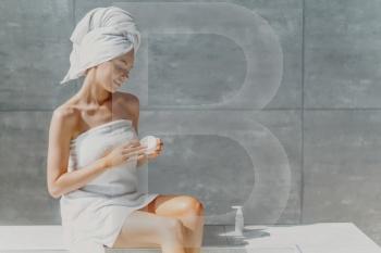 Horizontal shot of relaxed beautiful womam applies body lotion and nourishing cream after shower, poses in bathroom against grey wall, sits wrapped in towel, undergoes beauty treatments. Spa concept