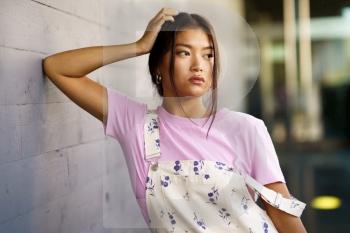 Young Asian woman in pink t shirt and stylish overalls touching head and looking away while leaning on gray wall on city street. Stylish Asian female leaning on wall