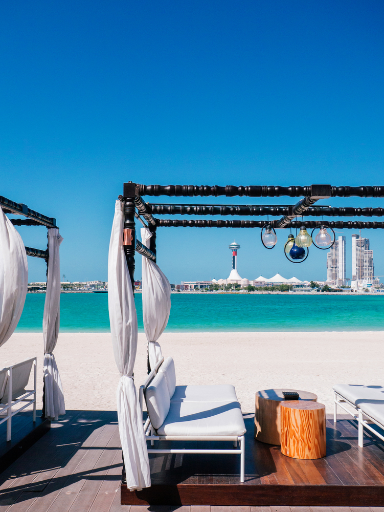 JAN 31, 2015 Abu Dhabu, UAE - Modern Beach Gazebo white curtain with white seat and wooden stool table. Blue sea under clear summer sky at Corniche beach with Marina island in background