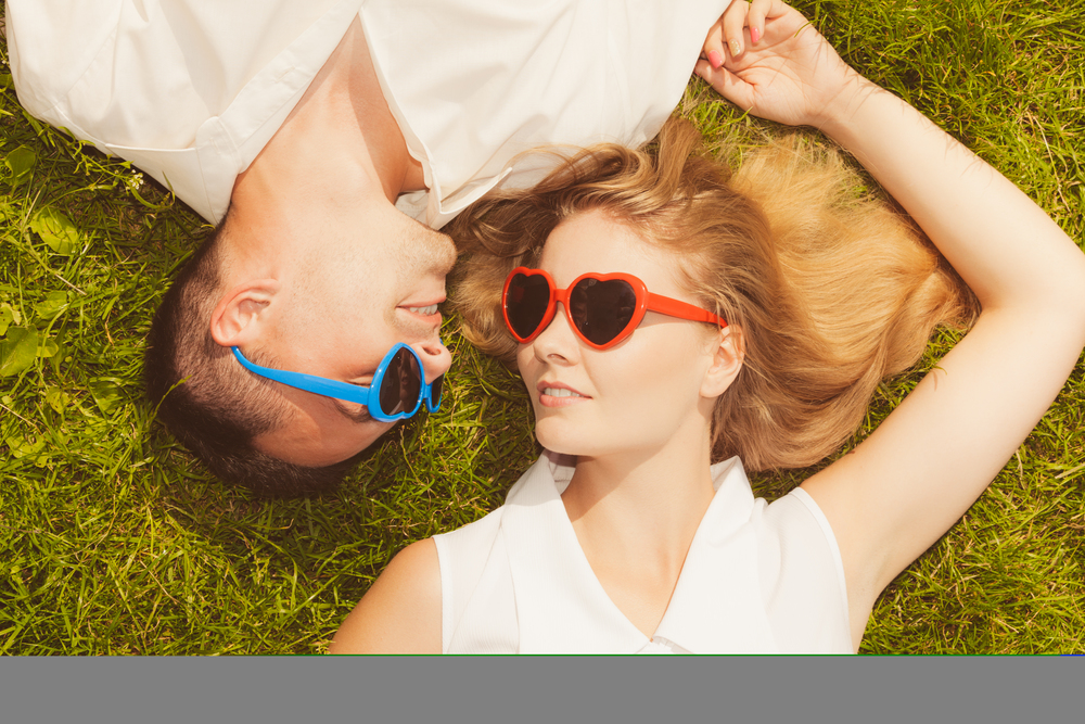 Love, relationship goals, funny accessories concept. Man and woman wearing heart shape sunglasses lying on grass, looking happy.. Man and woman wearing heart shape sunglasses