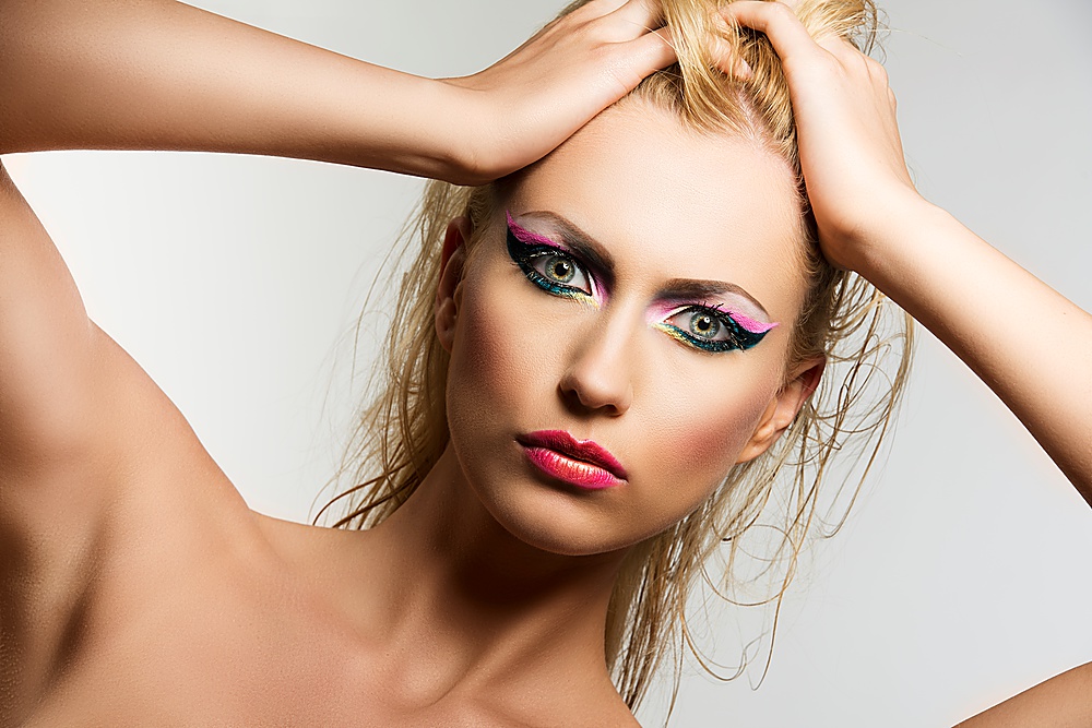 beautiful blonde girl with showy make up, her hands are on the head and she looks in to the lens