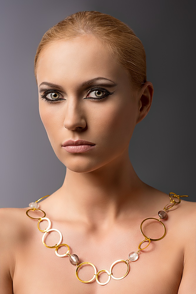 pretty blonde girl with naked shoulders, cute make-up and gold necklace with rings, looks in to the lens on gray background
