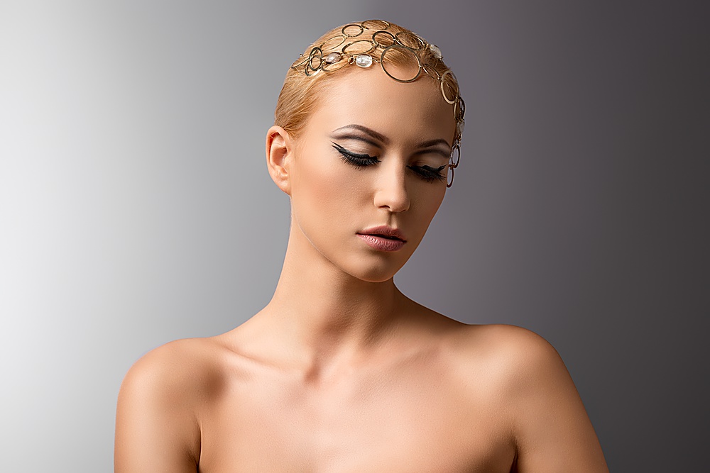 beautiful blonde girl with clean skin, nude shoulders and cute make-up wearing gold jewelry on the head. Gray background