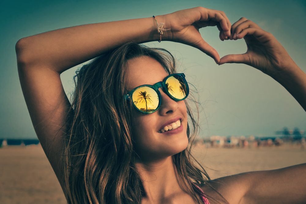 Brunette girl on beach sunglasses with palm tree reflection and fingers heart shape gesture