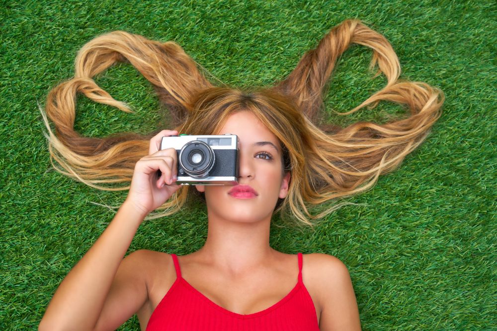 Blond teen girl with hair heart shapes lying down on turf with vintage photo camera