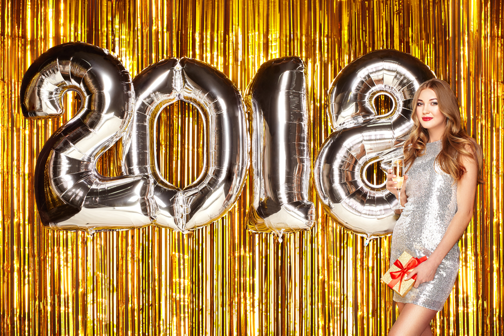 Beautiful Woman celebrating New Year. Girl with Glass of Champagne. Girl posing with Silver colored 2018 number Balloons over Golden sparkly background. Happy New Year