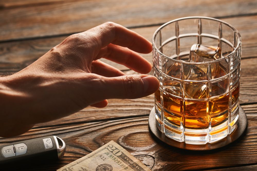 Man&rsquo;s hand reaching to glass of whiskey or alcohol drink with ice cubes and car key on rustic wooden table. Drink and drive and alcoholism concept. Safe and responsible driving concept.