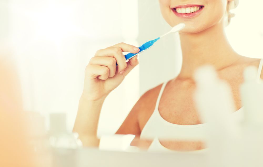 health care, dental hygiene, people and beauty concept - close up of smiling young woman with toothbrush cleaning teeth and looking to mirror at home bathroom. woman with toothbrush cleaning teeth at bathroom