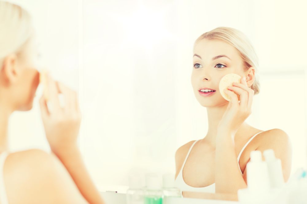 beauty, skin care and people concept - smiling young woman washing her face with facial cleansing sponge at bathroom. young woman washing face with sponge at bathroom
