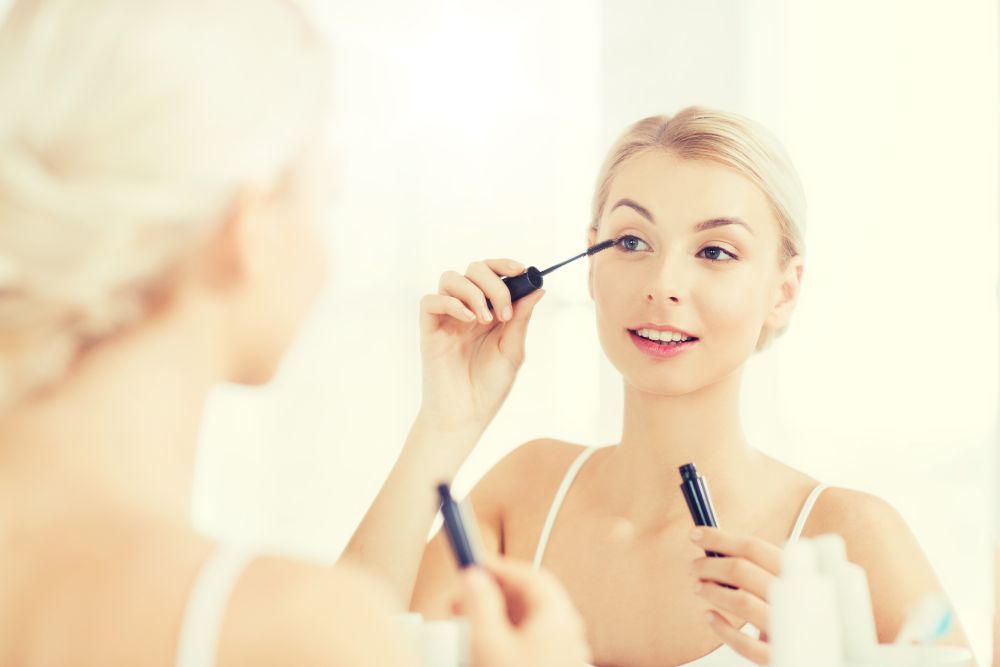 beauty, make up, cosmetics, morning and people concept - smiling young woman applying eye makeup with mascara and looking to mirror at home bathroom. woman with mascara applying make up at bathroom