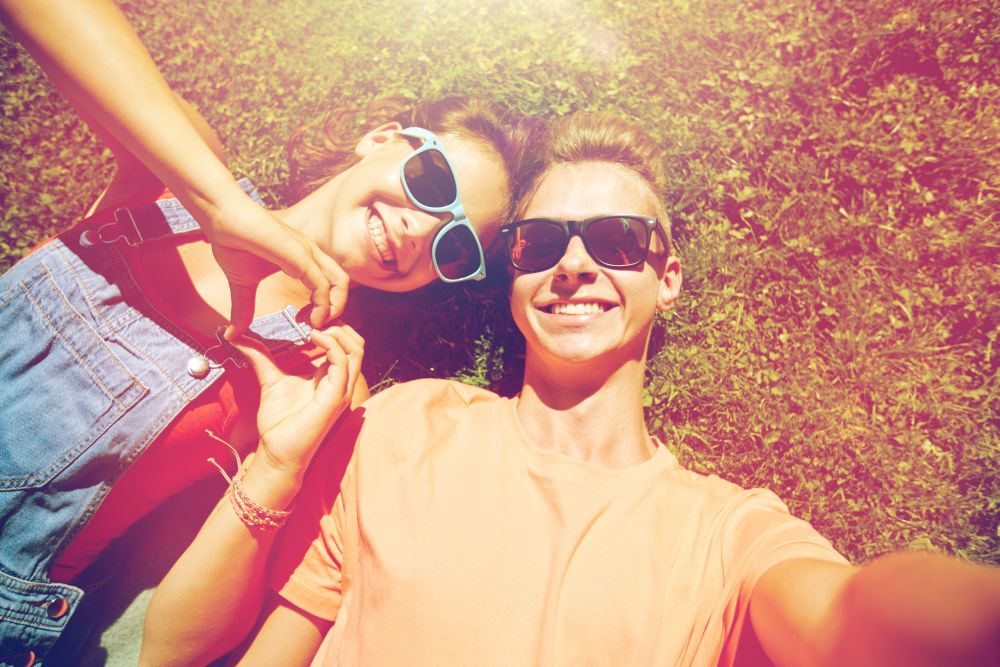 love and people concept - happy teenage couple in sunglasses lying on grass and taking selfie at summer. happy teenage couple taking selfie on summer grass