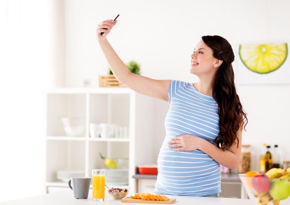pregnancy, people and technology concept - happy pregnant woman with smartphone taking selfie and having breakfast at home kitchen. happy pregnant woman with smartphone at kitchen