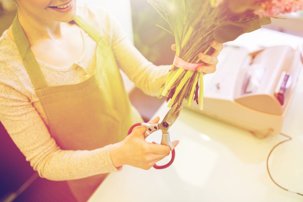 people, business, sale and floristry concept - close up of florist woman making bunch and cropping stems by scissors at flower shop. close up of woman with flowers and scissors