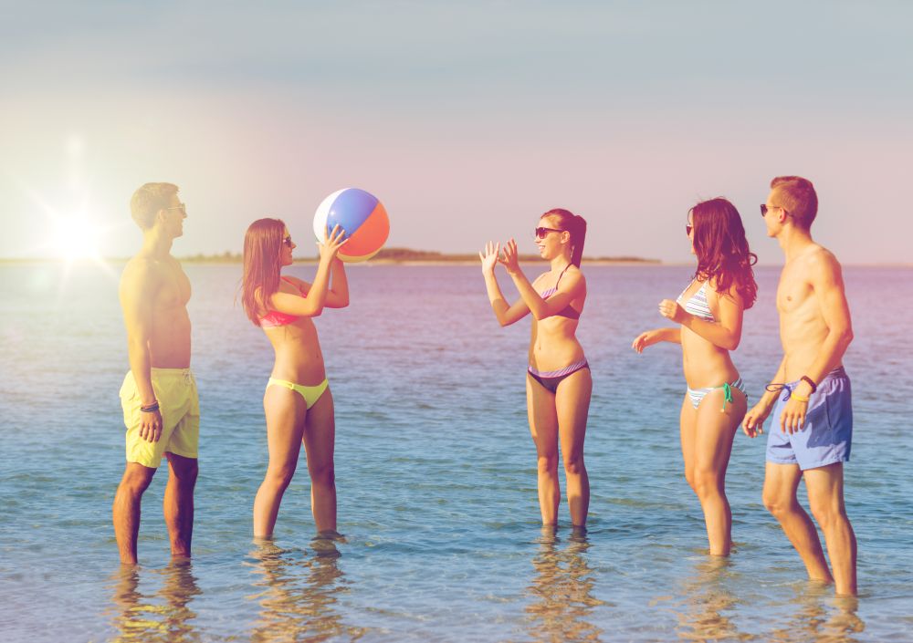 friendship, sea, summer vacation, holidays and people concept - group of smiling friends wearing swimwear and sunglasses playing with inflatable ball on beach. smiling friends in sunglasses on summer beach