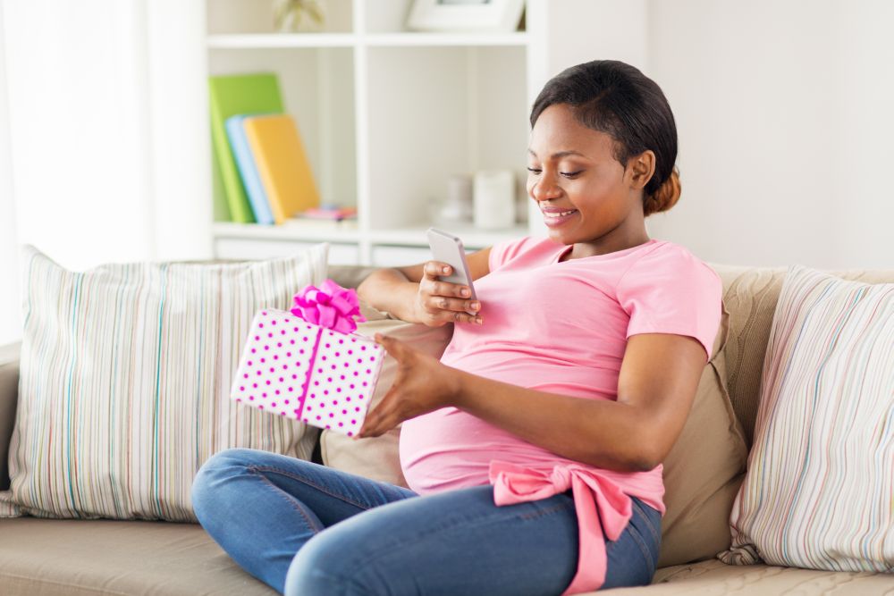 pregnancy, holidays and people concept - happy pregnant african american woman sitting on sofa with smartphone and gift box at home. african pregnant woman with smartphone and gift