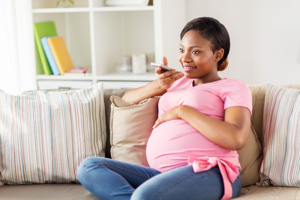 pregnancy, technology and people concept - happy pregnant african american woman using voice command recorder or calling on smartphone at home. pregnant woman using voice recorder on smartphone