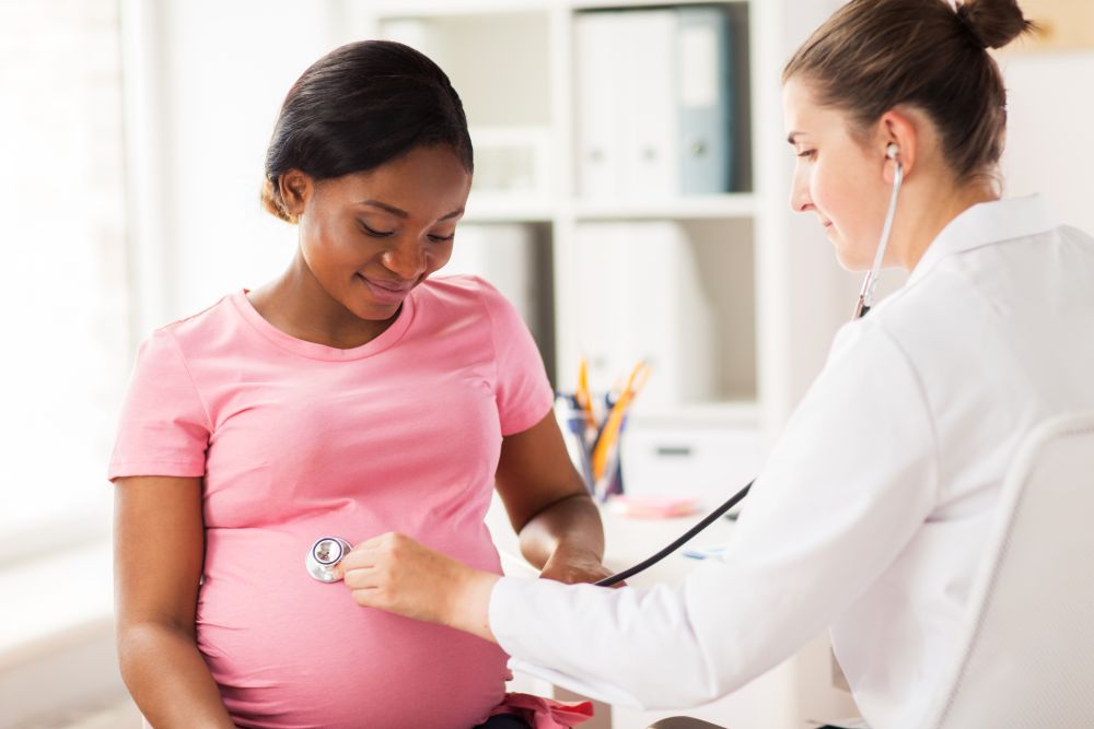 pregnancy, medicine, healthcare and people concept - gynecologist doctor with stethoscope listening to pregnant african american woman baby heartbeat at hospital. gynecologist doctor and pregnant woman at hospital