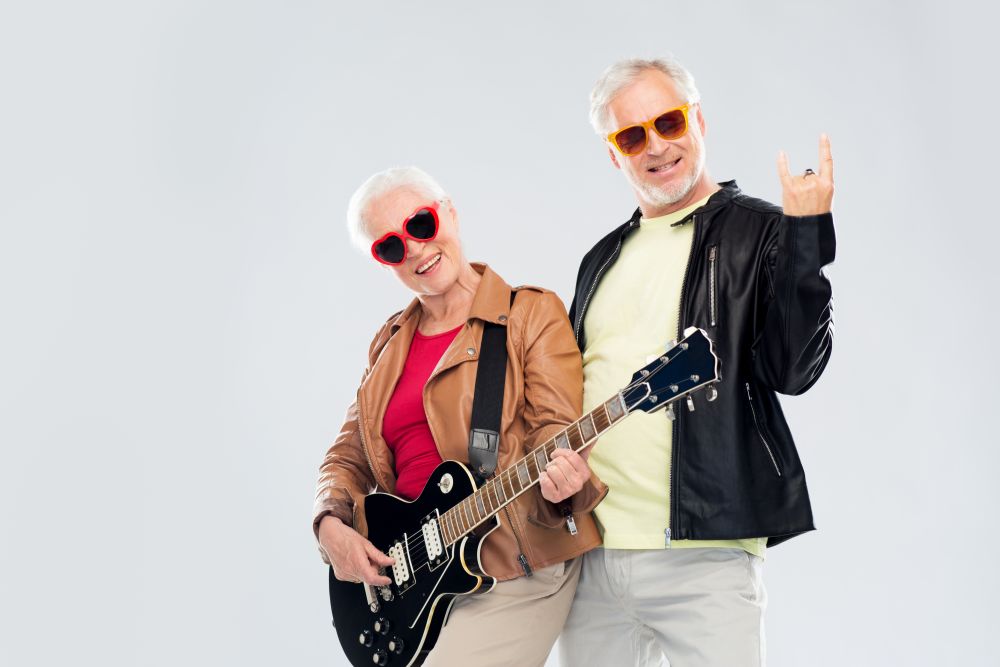 music, age and people concept - happy senior couple in sunglasses with electric guitar showing rock hand sign. senior couple with guitar showing rock hand sign