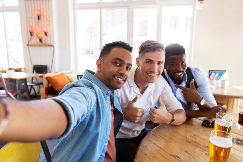 people, friendship and leisure concept - happy male friends drinking beer, taking selfie and showing thumbs up at bar or pub. friends taking selfie and drinking beer at bar