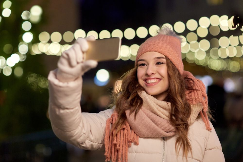 holidays and people concept - beautiful happy young woman taking selfie over christmas lights in winter evening. young woman taking selfie over christmas tree