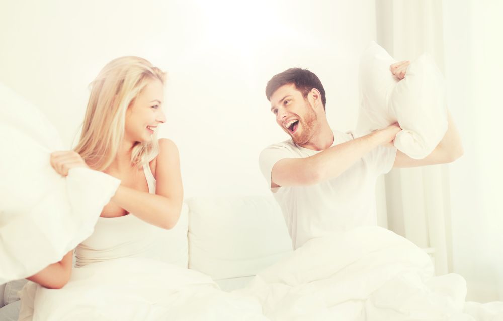 people, family, fun, bedtime and fun concept - happy couple having pillow fight in bed at home. happy couple having pillow fight in bed at home