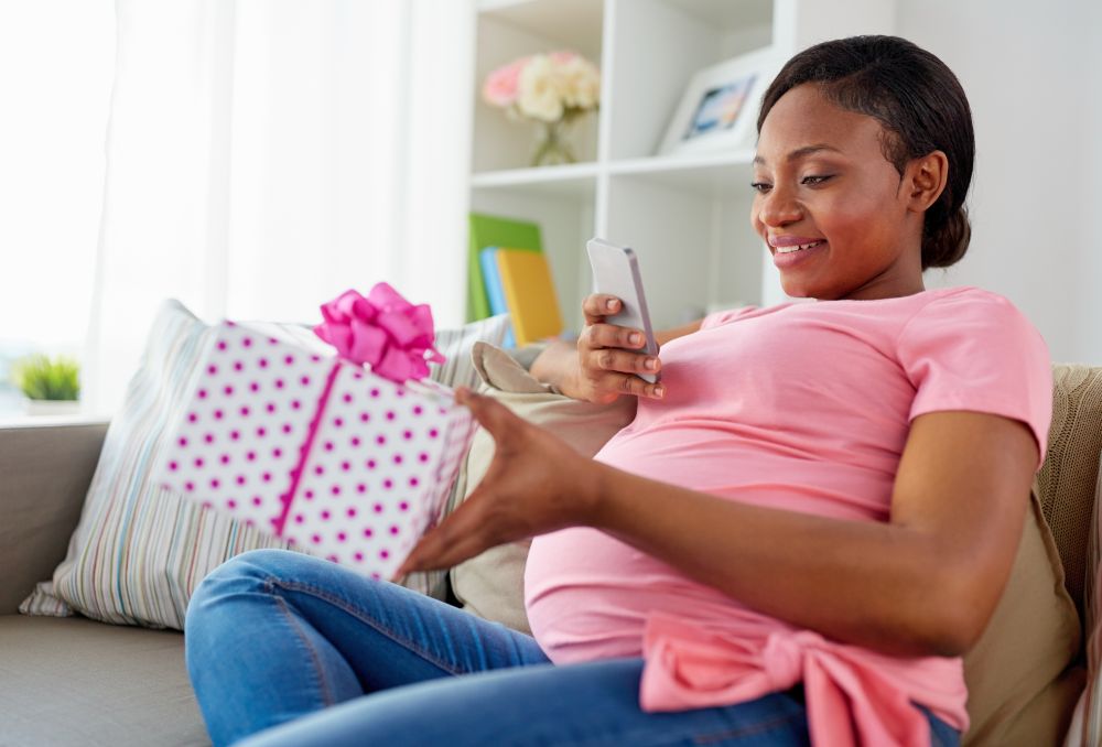 holidays and technology concept - happy pregnant african american woman sitting on sofa at home and taking picture of gift box with smartphone. african pregnant woman with smartphone and gift