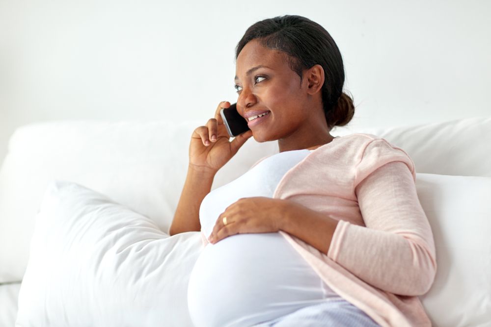 pregnancy, technology and people concept - happy pregnant african american woman calling on smartphone in bed at home. happy pregnant woman calling on smartphone at home