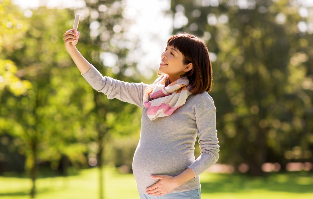 pregnancy, people and technology concept - happy pregnant asian woman with smartphone taking selfie at park. happy pregnant asian woman taking selfie at park