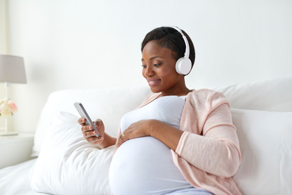 pregnancy, technology and people concept - happy pregnant african american woman with headphones and smartphone in bed at home. pregnant woman in headphones with smartphone