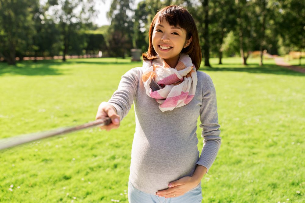 pregnancy, people and technology concept - happy pregnant asian woman with selfie stick taking picture at park. happy pregnant asian woman taking selfie at park