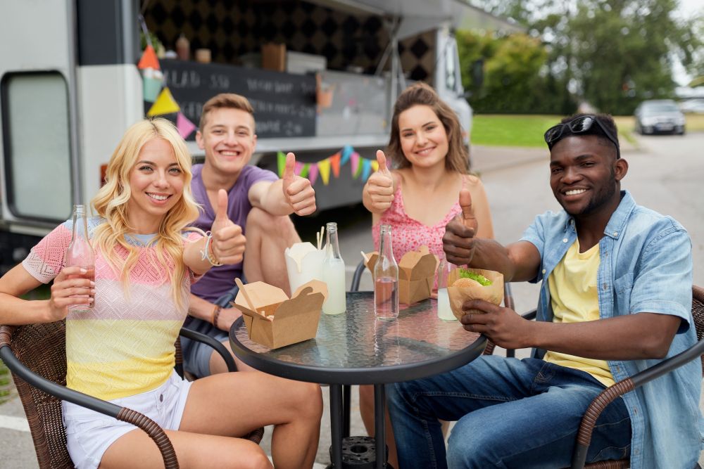 leisure and people concept - happy friends with drinks eating at food truck showing thumbs up. happy friends with drinks eating at food truck