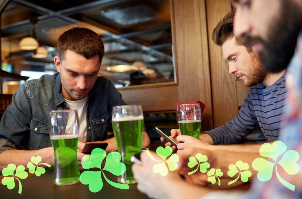 st patricks day, technology and leisure concept - male friends drinking green beer and messaging on smarphones at bar or pub. friends with smarphones and green beer at pub