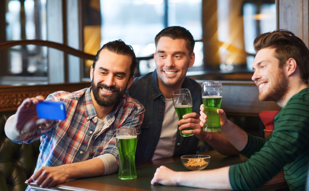 st patricks day, leisure and technology concept - happy male friends drinking green beer and taking picture with smartphone selfie stick at bar or pub. friends taking selfie with green beer at pub