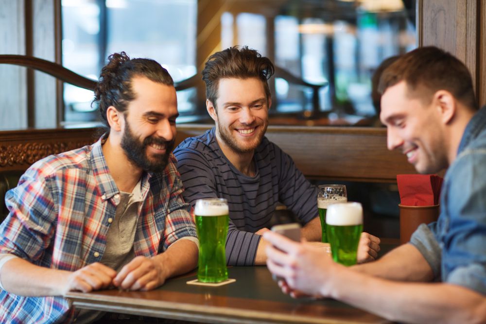 leisure, st patricks day and technology concept - happy male friends with smartphone drinking green beer at bar or pub. friends with smartphone drinking green beer at pub