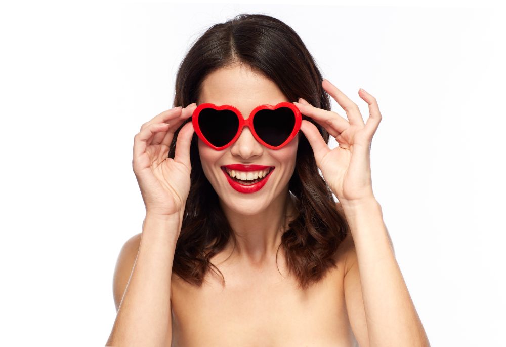 valentines day, beauty and people concept - happy smiling young woman with red lipstick and heart shaped sunglasses over white background. woman with red lipstick and heart shaped shades
