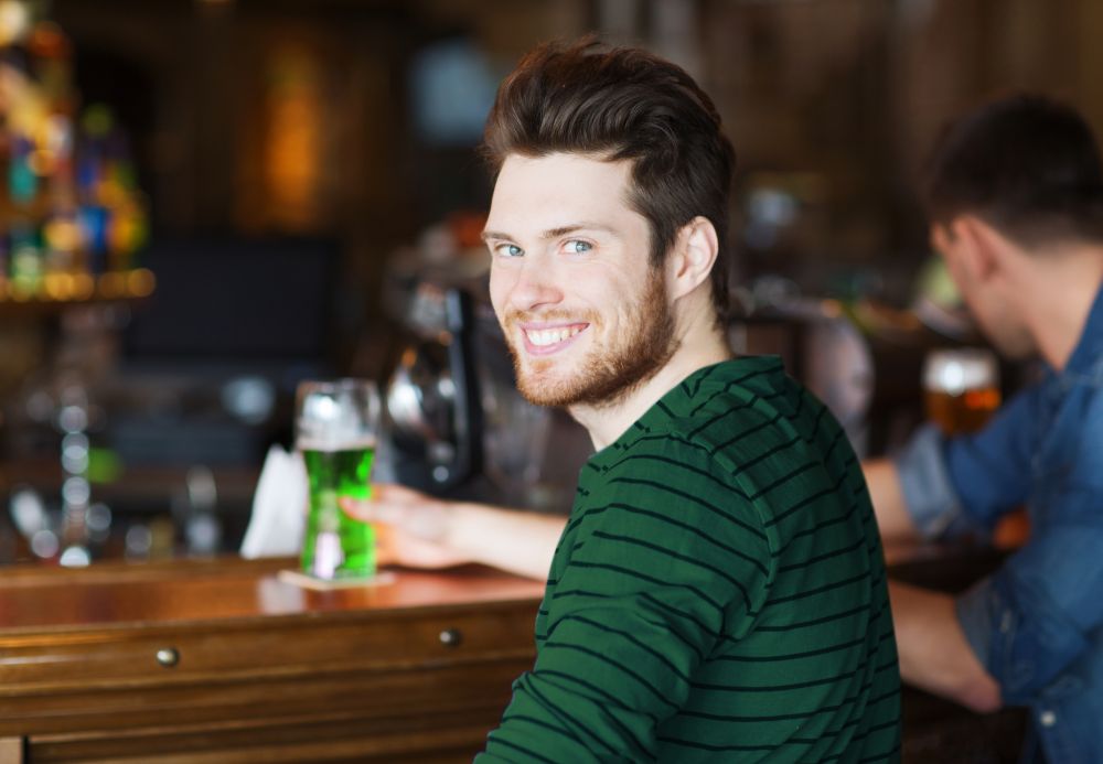 people, leisure and st patricks day concept - happy smiling young man drinking green beer at bar or pub. happy man drinking green beer at bar or pub