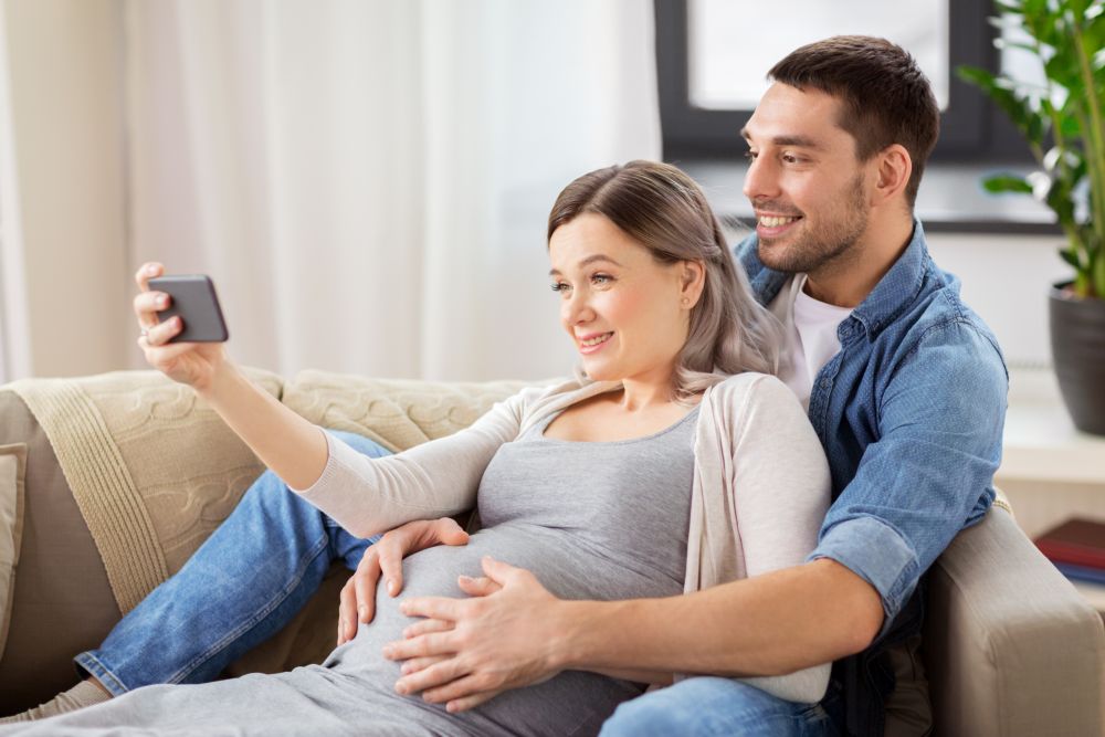 technology, pregnancy and people concept - happy man and his pregnant wife taking selfie by smartphone at home. man and pregnant woman taking selfie at home