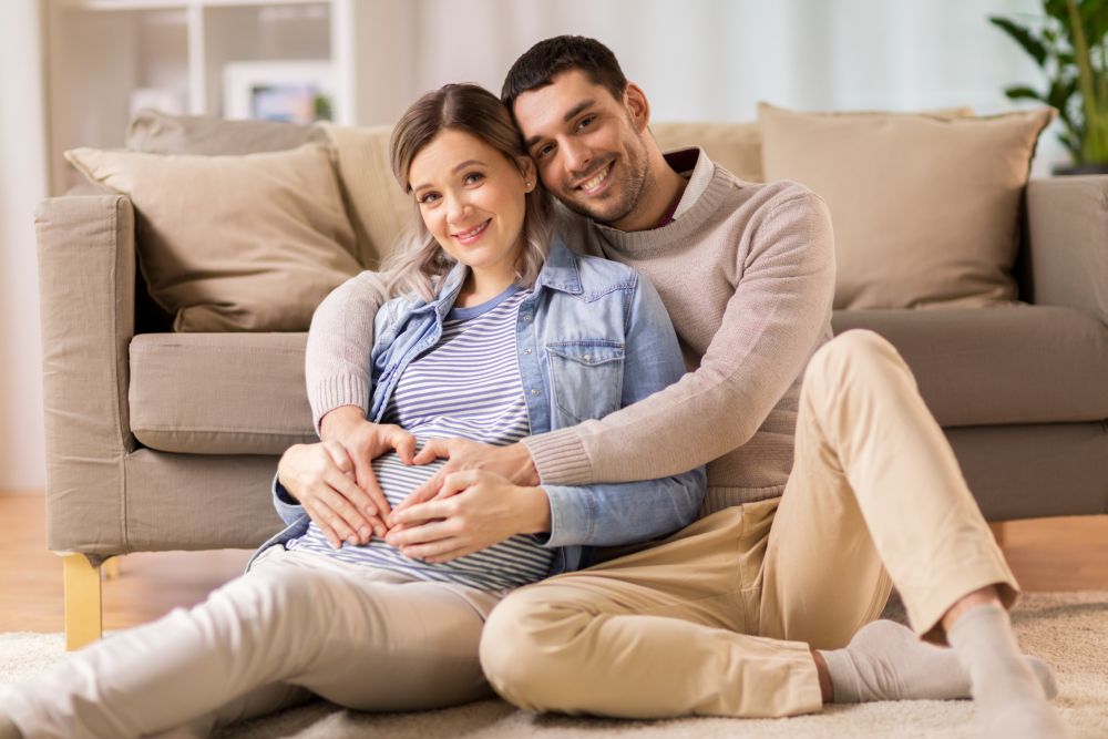 pregnancy, love and care concept - happy man hugging pregnant woman and making hand heart gesture at home. man with pregnant woman making hand heart at home