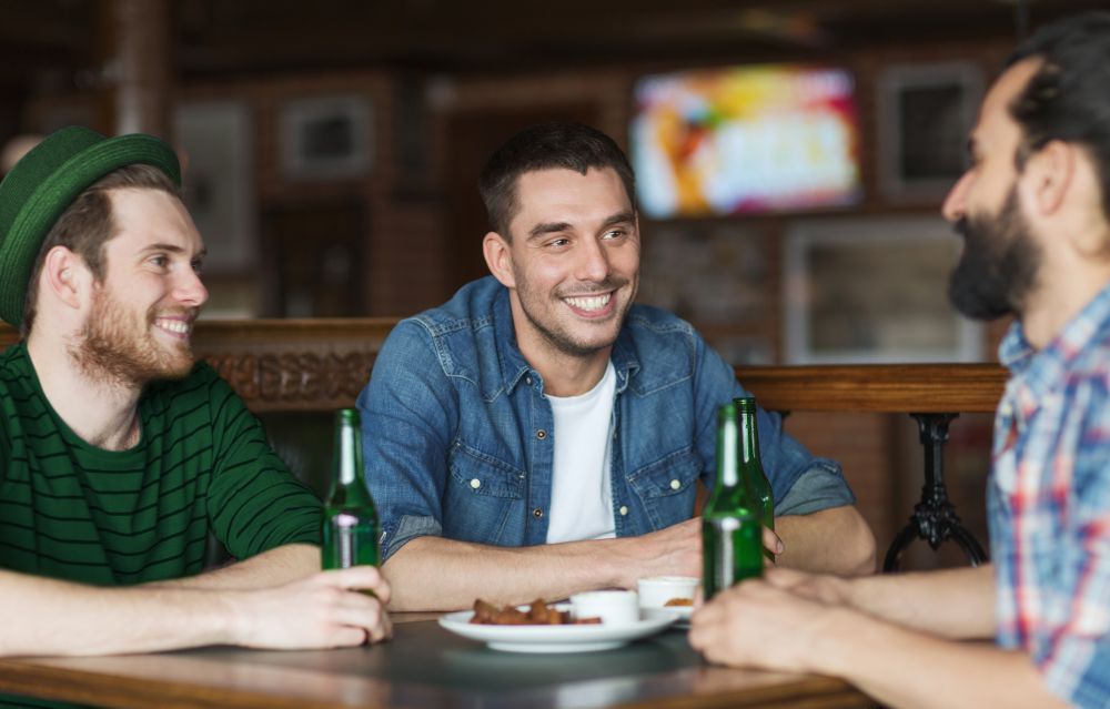 people, leisure, alcohol and st patricks day concept - happy male friends drinking bottled beer and talking at bar or pub. happy male friends drinking beer at bar or pub