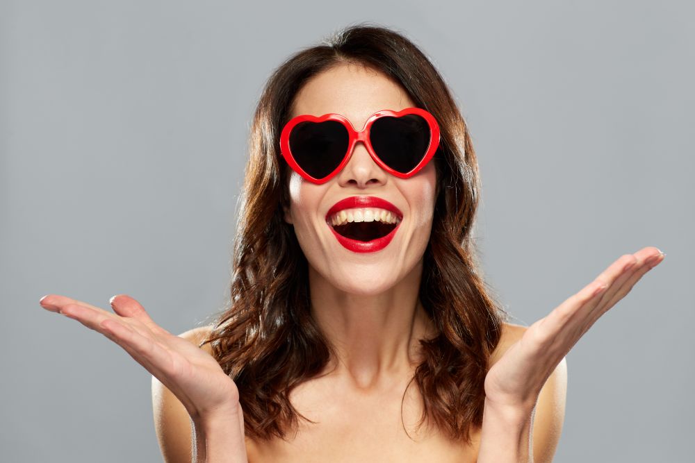 valentines day, beauty and people concept - happy smiling young woman with red lipstick and heart shaped sunglasses over gray background. woman with red lipstick and heart shaped shades