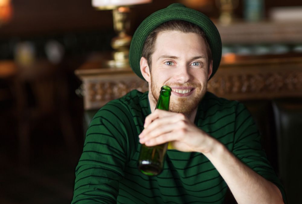 people, leisure and st patricks day concept - portrait of happy young man in green hat drinking beer from bottle at bar or pub. man drinking beer from green bottle at bar or pub