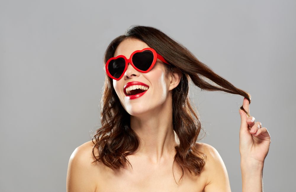 valentines day, beauty and people concept - happy smiling young woman with red lipstick and heart shaped sunglasses curling hair strand over white background. woman with red lipstick and heart shaped shades