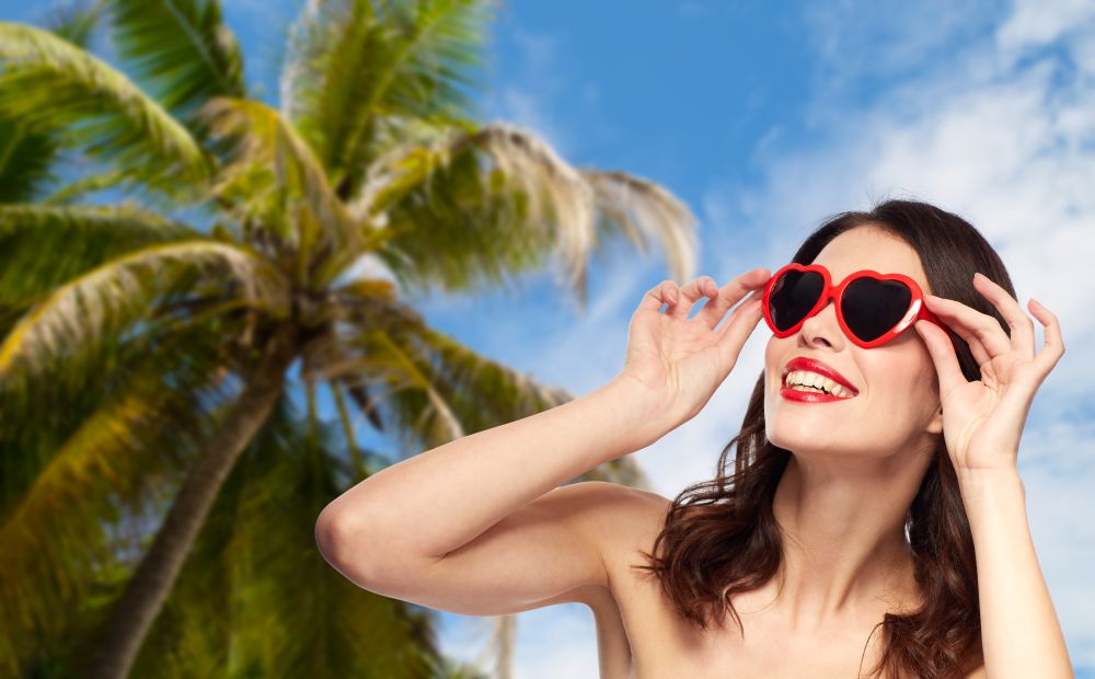 travel, tourism, summer vacation and valentines day concept - happy smiling young woman with red lipstick and heart shaped sunglasses over palm tree and blue sky background. woman with red lipstick and heart shaped shades