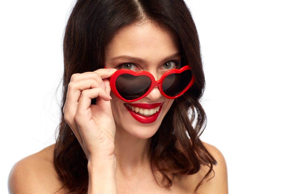 valentines day, beauty and people concept - close up of happy smiling young woman with red lipstick and heart shaped sunglasses over white background. woman with red lipstick and heart shaped shades
