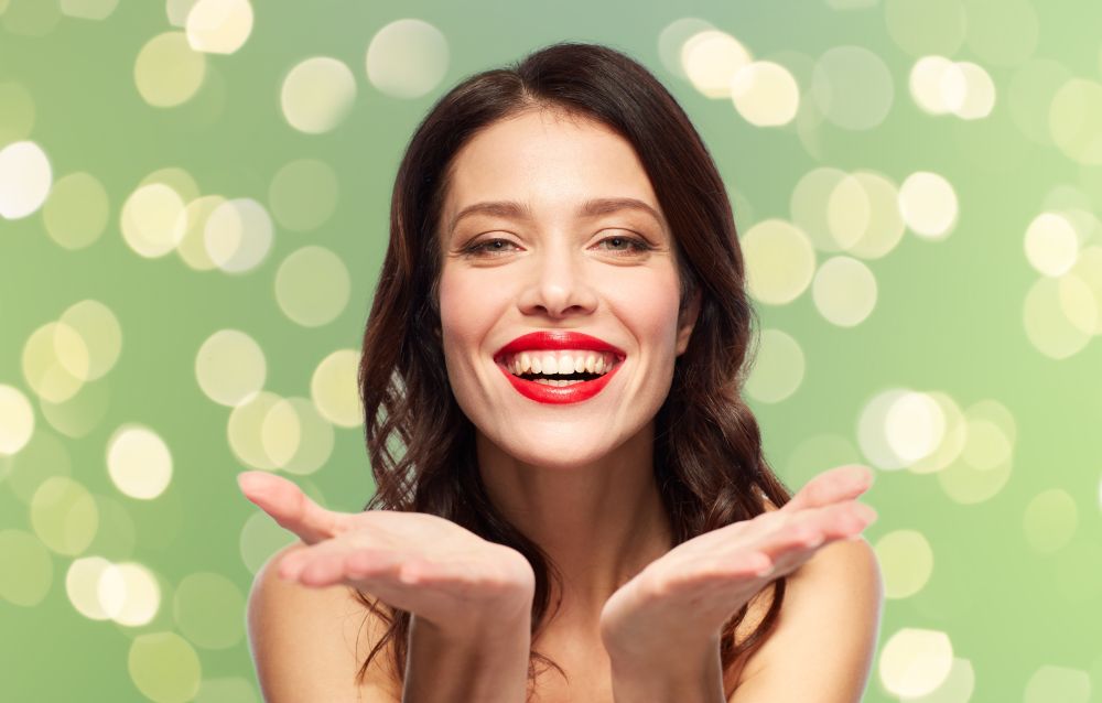 beauty, make up and people concept - happy smiling young woman with red lipstick holding something imaginary on palms over summer green lights background. beautiful smiling young woman with red lipstick