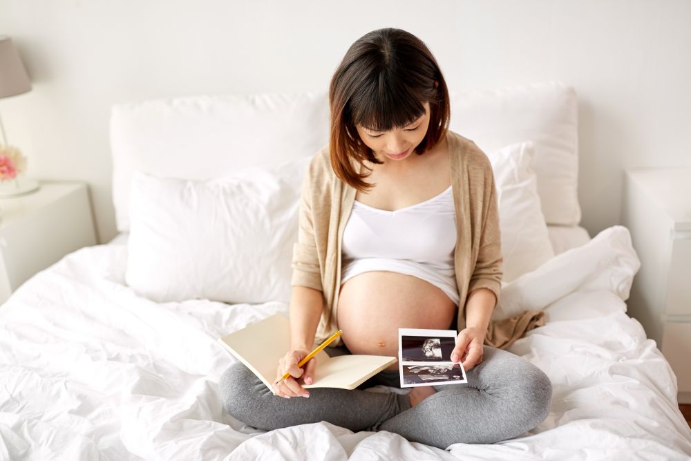 pregnancy and people concept - happy pregnant asian woman with fetal ultrasound image and notebook or diary in bed at home. pregnant woman with fetal ultrasound image at home