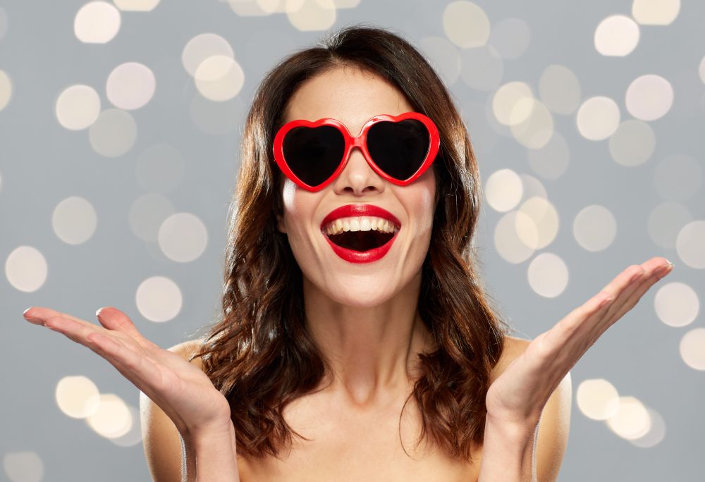 valentines day, holidays, beauty and people concept - happy smiling young woman with red lipstick and heart shaped sunglasses over gray background and lights. woman with red lipstick and heart shaped shades