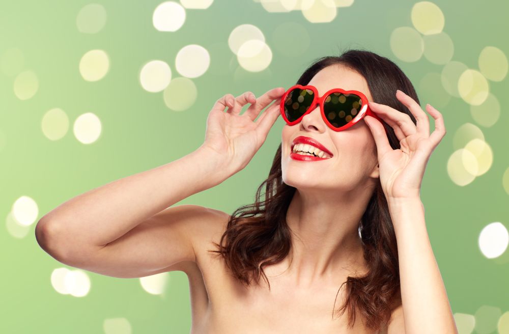 valentines day, beauty and people concept - happy smiling young woman with red lipstick and heart shaped sunglasses over green background with lights. woman with red lipstick and heart shaped shades
