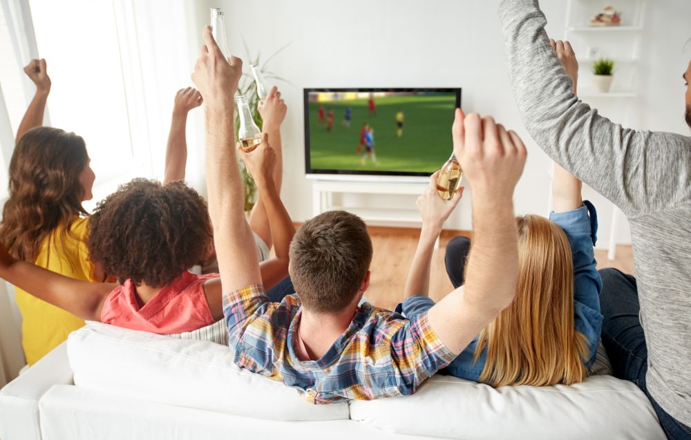 friendship, leisure and people concept - happy friends with non-alcoholic beer sitting on sofa and watching soccer or football game on tv at home. friends with beer watching soccer on tv at home
